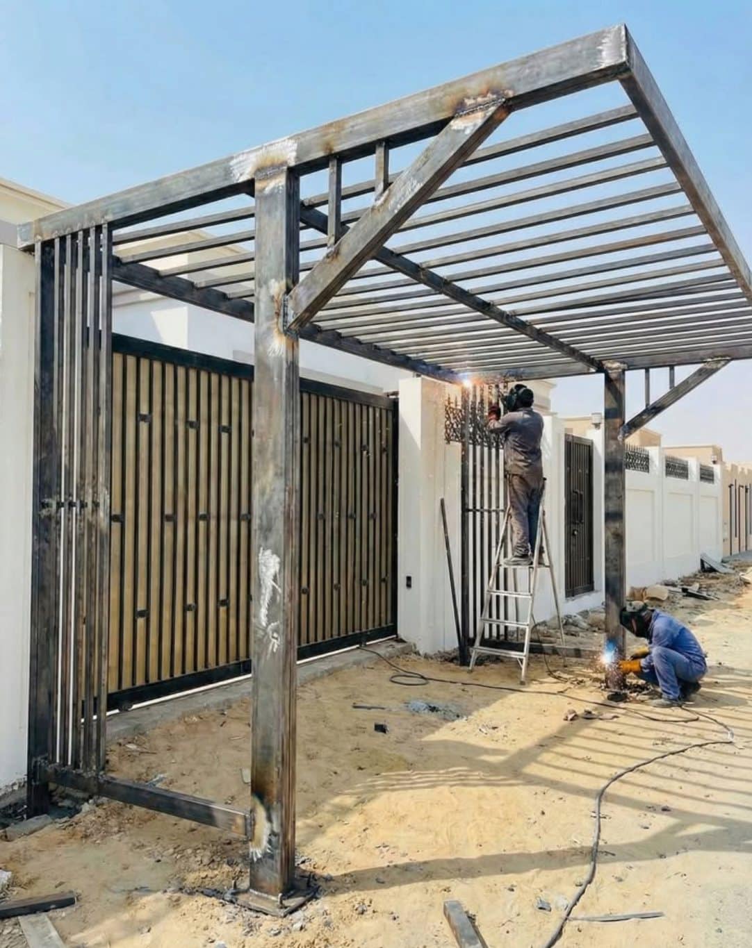 Pergola in ruw staal in montage voor een woning, twee lassers aan het werk aan de structuur en de poort