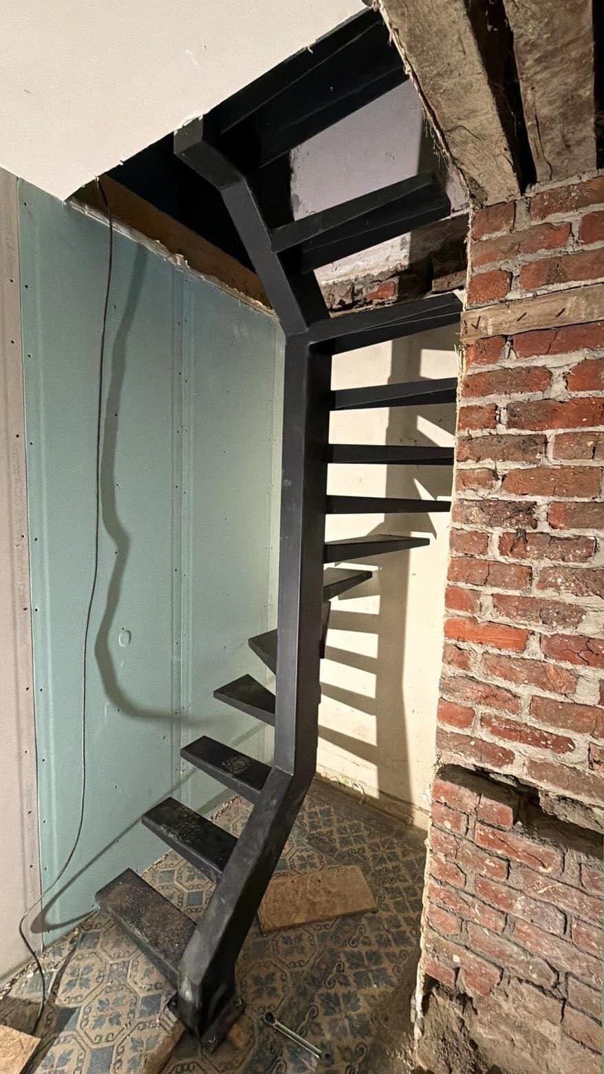 Zwarte stalen draaitrap met centrale trapboom, geplaatst tegen een oude bakstenen muur in een woning in renovatie
